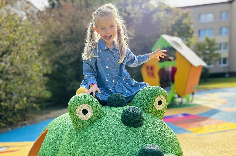 A young girl in a blue dress sits and smiles on top of a green frog-shaped playground structure outdoors, highlighting the safety and fun offered by rubber surfacing applications for playgrounds.