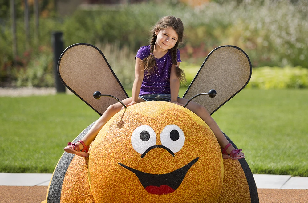 A young girl with braided hair sits on a large, smiling bee playground structure outdoors on a sunny day, enjoying the safe, colorful rubber surfacing applications for playgrounds beneath her.