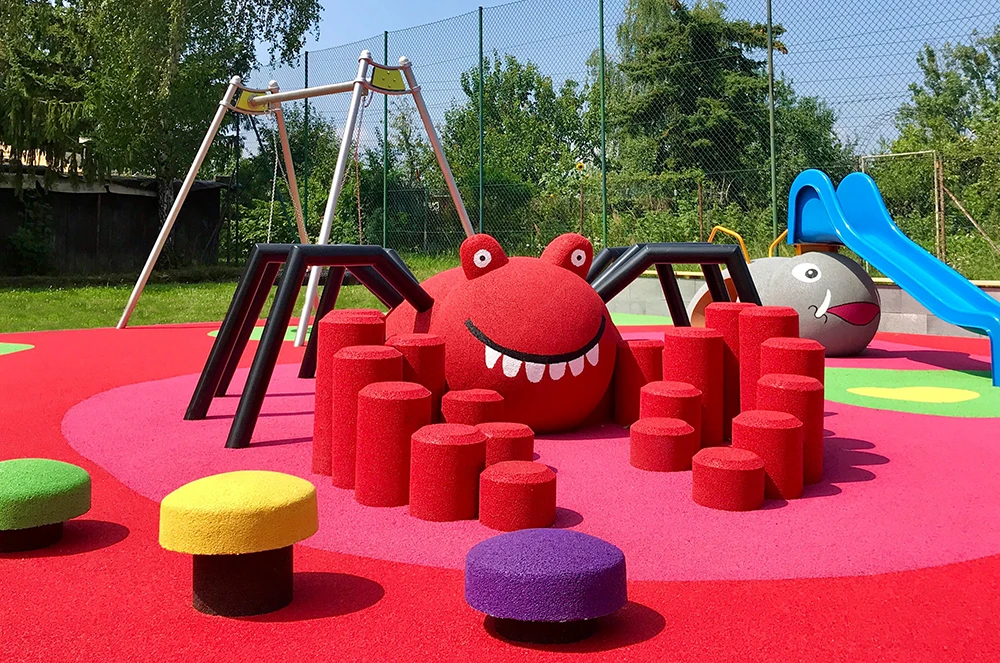 Outdoor playground with colorful, animal-themed play structures, including a red spider, swings, slide, and soft climbing blocks on bright rubber surfacing—ideal for playgrounds and commercial spaces. Trees and fence in the background.