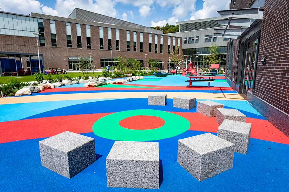 Colorful school playground with geometric patterns and rubber surfacing applications for playgrounds, square stone seats arranged in a circle, picnic table, outdoor play equipment, and a brick building in the background under a partly cloudy sky.
