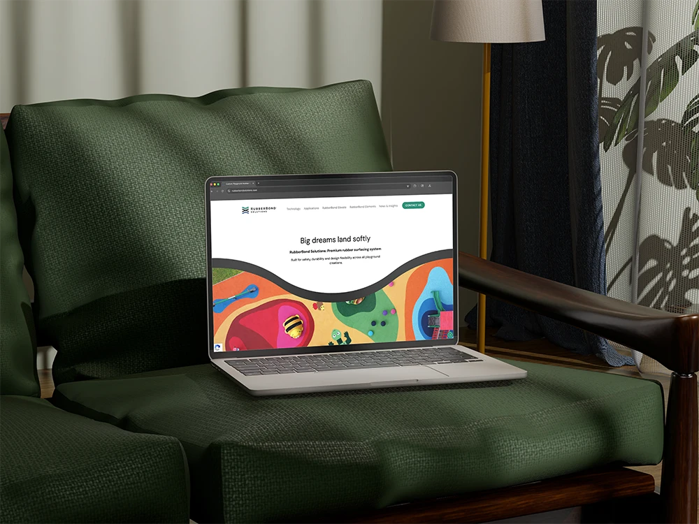 A laptop displaying a colorful website about rubber surfacing applications for playgrounds and commercial spaces sits on a green cushioned armchair in a well-lit room.