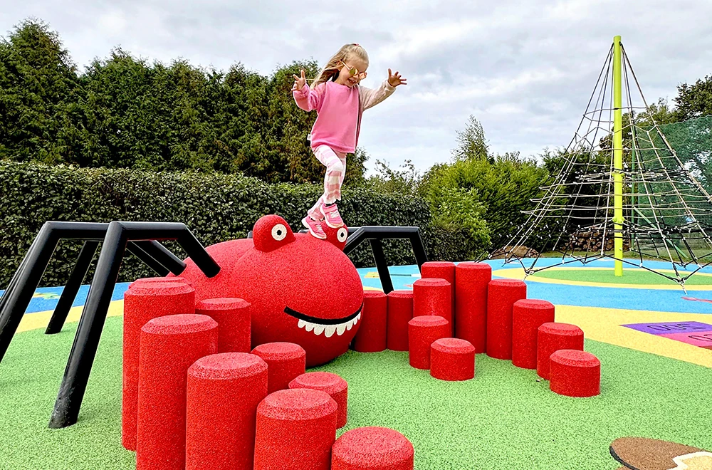 A young child in pink clothing balances on top of a large red spider-shaped playground structure with red stepping stones, showcasing the safety benefits of rubber surfacing applications for playgrounds and commercial spaces.