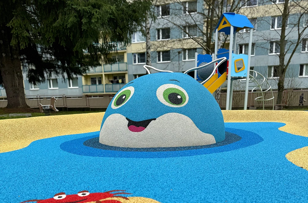 A playground structure shaped like a blue, smiling animal with green eyes and white ears stands on vibrant rubber surfacing applications for playgrounds, with apartment buildings rising in the background.